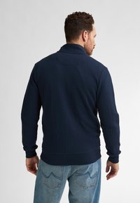 Námořnický modrý svetr s texturovaným vlnitým vzorem, má vysoký límec a pružné manžety, kombinovaný se světle modrými džíny.