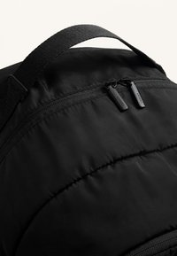 Fekete párnázott hátizsák sima, nylonszerű textúrával, dupla cipzárral és megerősített felső fogantyúval a könnyű hordozhatóság érdekében.