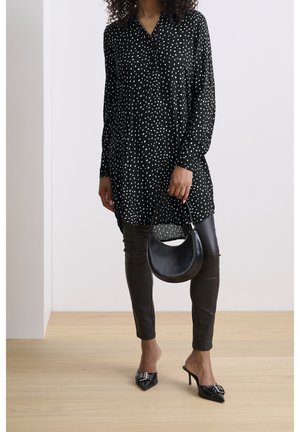 Woman wearing black and white spotted tunic, black leather leggings, black pointed heels, holding black crescent handbag.