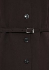 Pruun kleit, millel on põimitud talje ja vööga disain. Omaduseks on mustad nupud ja tekstuuriga kangas, mis loob vooluva silueti.