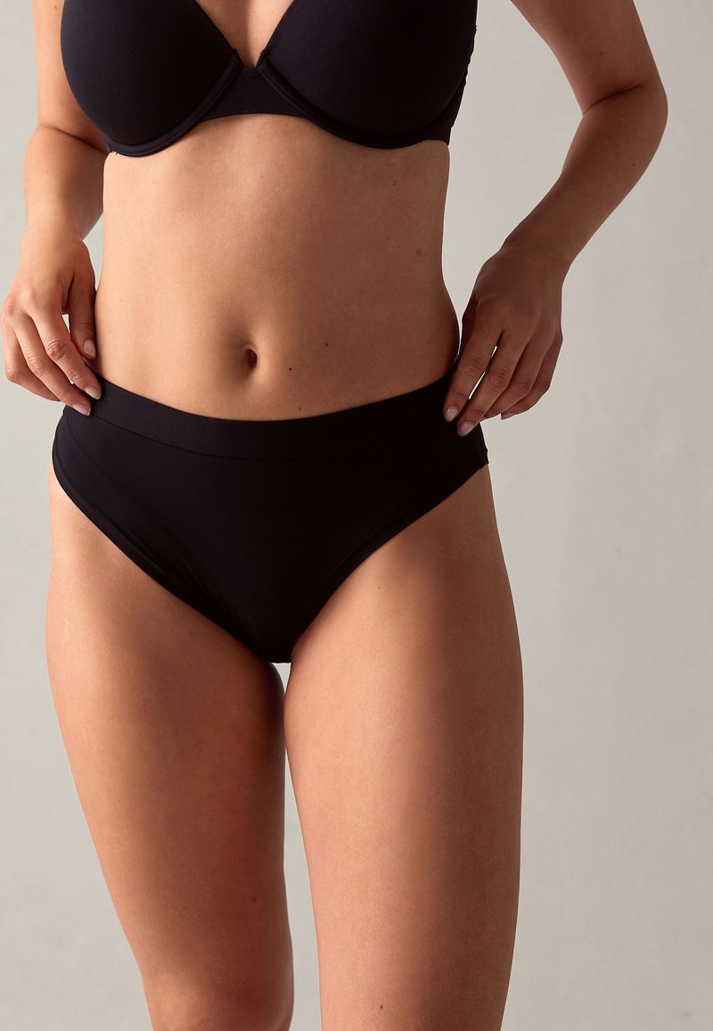 Mujer con sujetador negro y ropa interior negra de cintura alta, manos descansando cerca de las caderas, sobre fondo neutro.
