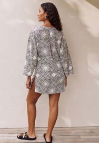 Black and white patterned tunic with long sleeves, featuring geometric designs and side slits, paired with black sandals on a wooden surface.