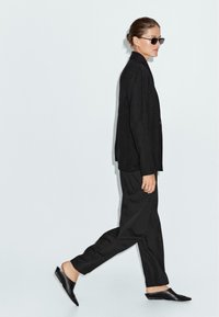 Blazer nero oversize, pantaloni neri larghi e ballerine nere appuntite. Gli accessori includono occhiali da sole scuri. Modella che cammina su uno sfondo chiaro.
