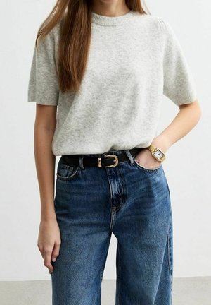 Kvinde iført kortærmet, lysegrå sweater, højtaljede blå jeans med sort bælte, guld armbåndsur, hånd i lommen.