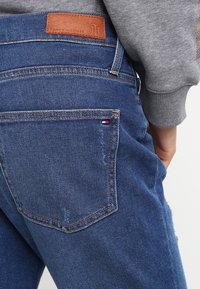 Siniset farkut, joissa on nahkainen logo-patch vyötäröllä, kontrastiväriset ompeleet ja takatasku, jossa on pieni logo-yksityiskohta.