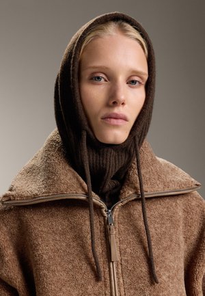 Tânără cu părul deschis la culoare, purtând o cagula din tricot maro și o jachetă din polar căptușită maro, cu fermoar, pe un fundal neutru.