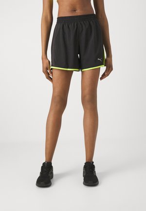 Pantalones cortos deportivos negros con detalles en verde neón, cintura elástica y un material ligero. Combinados con zapatos deportivos negros.