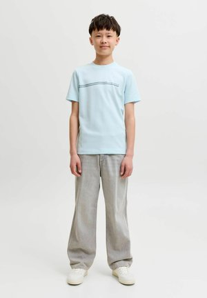 Garçon debout de face portant un t-shirt bleu clair, un jean large gris et des baskets blanches sur un fond clair uni.