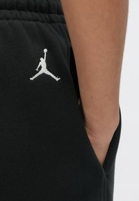 Κοντινό πλάνο χεριού ατόμου σε τσέπη μαύρων παντελονιών με λευκό λογότυπο Jumpman κεντημένο κοντά στη μέση.