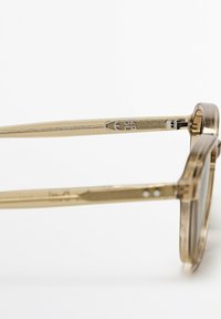 Genomskinlig beige glasögonram med rektangulär form. Metallackenter på bågarna, ingraverade detaljer och små hål nära gångjärnen.