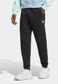 Black sweatpants with a tapered fit, featuring side pockets and an embroidered white logo on the left thigh, paired with white sneakers.