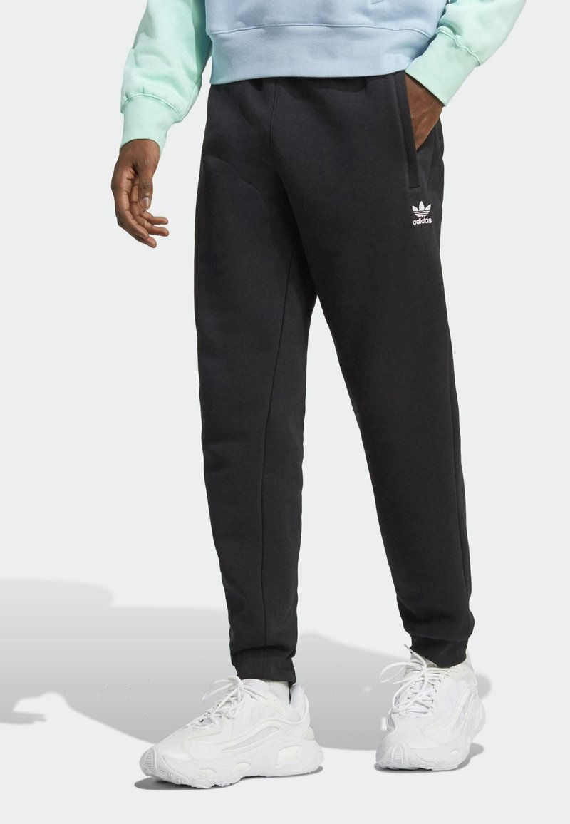Black sweatpants with a tapered fit, featuring side pockets and an embroidered white logo on the left thigh, paired with white sneakers.