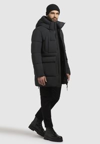 Black puffer jacket with a hood, featuring a front zipper, multiple pockets, and a quilted texture. Paired with black pants and boots.