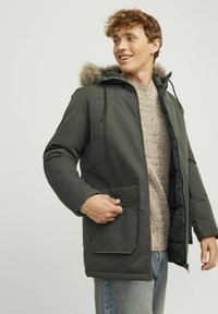 Donkergroene parka met een met bont gevoerde capuchon, voorkeurszakken en ritssluiting. Gedragen over een gestructureerde crèmekleurige trui.