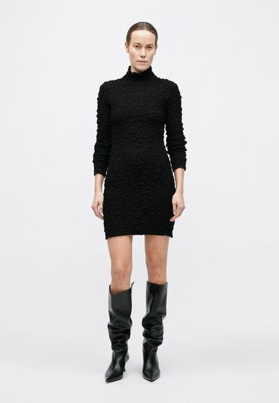 Naine seisab, seljas tekstureeritud must pika varrukaga minikleidi ja mustad kotis põlveni ulatuvad saapad, taust on ühtlaselt valge.