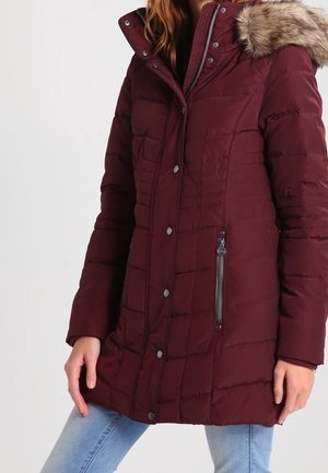 Kobieta ubrana w bordowy pikowany zimowy płaszcz z kapturem obszytym futrem i zasuwanymi kieszeniami po bokach, w połączeniu z jasnoniebieskimi dżinsami.