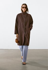 Brun, lätt oversized trenchcoat med hög krage, framknappar och sidofickor, i kombination med ljusblå jeans och sandaler.