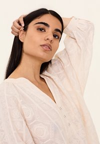 Light pink blouse with a textured pattern, V-neckline, and button details. Long sleeves with gathered cuffs. Soft fabric.