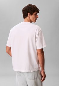 Ανδρας με κοντά καστανά σγουρά μαλλιά, που φοράει απλό λευκό φαρδύ κοντομάνικο μπλουζάκι και ανοιχτό γκρι τζιν, με την πλάτη στραμμένη προς ουδέτερο φόντο.