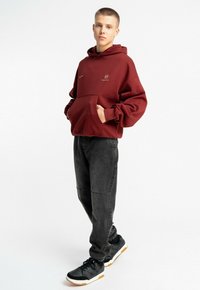 Bordowa bluza z kapturem z kieszenią kangurką, czarne dżinsy oraz czarne sneakersy z białą i gumową podeszwą. Bluza posiada małe logo na piersi.