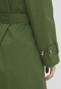Olive krāsas garā mēteli ar teksturētu audumu, ar jostu viduklī un pogām uz aprocēm. Iespējas eleganta atpakaļšuvuma detaļa.