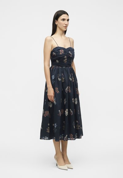 Femme aux longs cheveux foncés portant une robe midi sans manches bleu marine avec une broderie florale colorée et des talons beige à bout pointu, debout de profil.