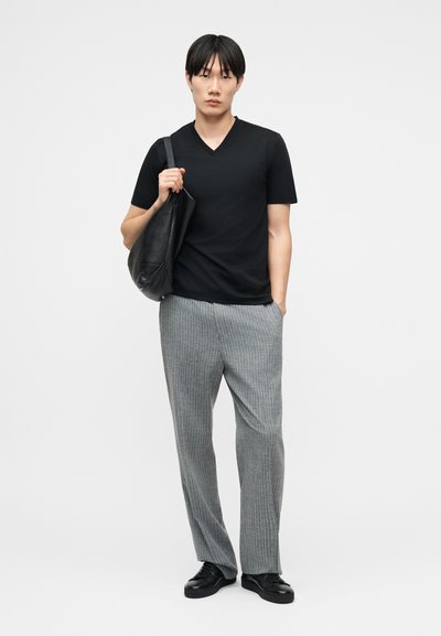 Νεαρός άνδρας στέκεται με το ένα χέρι στην τσέπη, φοράει μαύρο πουκάμισο με V λαιμόκοψη, γκρι παντελόνι με λεπτές ρίγες, μαύρα παπούτσια και κρατάει μία μαύρη δερμάτινη τσάντα στον ώμο.