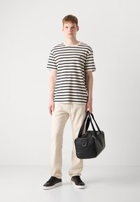 Black leather duffel bag with dual handles, textured finish, and metal hardware. Model wears a striped T-shirt, beige pants, and black sneakers.