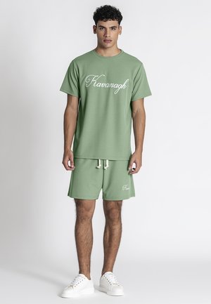 Uomo in piedi che indossa una maglietta Kavanagh color salvia e pantaloncini con sneakers bianche su uno sfondo chiaro e uniforme.