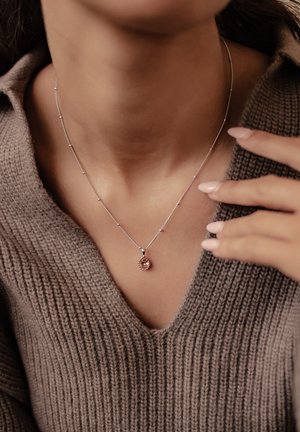 Silver necklace with a teardrop pendant featuring a pink stone, resting on a v-neck sweater with a ribbed texture.