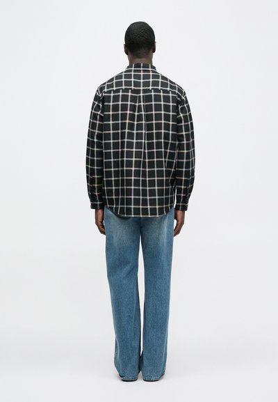 Μαύρο και άσπρο πουκάμισο με καρό μοτίβο, μακριά μανίκια και κυλινδρική εφαρμογή, συνδυασμένο με άνετο μπλε τζιν παντελόνι. Το φόντο είναι απλό λευκό.