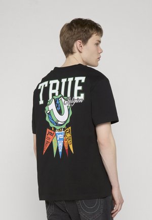 Junger Mann im Seitenprofil, trägt ein schwarzes T-Shirt mit der Aufschrift "TRUE" und bunten Stadtwimpeln auf der Rückseite vor einem schlichten Hintergrund.