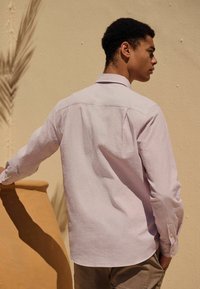 Světle růžová strukturovaná košile s límečkem, dlouhými rukávy a knoflíkovými manžetami. Obývá ji osoba, přičemž důraz je kladen na záda a rameno.