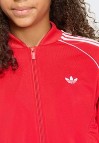 Κόκκινο μπουφάν με φερμουάρ, με λεία υφή, που διαθέτει λευκές ρίγες στους ώμους και λευκό λογότυπο της Adidas στο αριστερό στήθος.