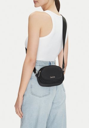 Donna che indossa jeans azzurri chiari e top bianco senza maniche con borsa a tracolla nera DeeZee appoggiata sulla spalla.