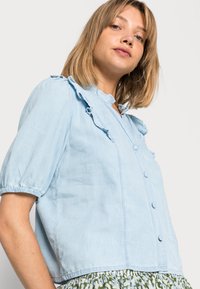 Blusa de denim azul claro, com mangas curtas e detalhes em renda ao longo do colarinho e fecho com botões; tecido texturizado e corte ligeiramente largo.