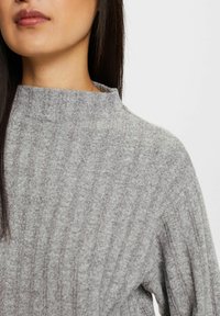 Harmaa ribbikuvioinen neule, jossa on korkea kaulus, esittelee pehmeää tekstuuria ja tasaista ommelta. Kankaassa on hienovarainen kiilto ja rento istuvuus.