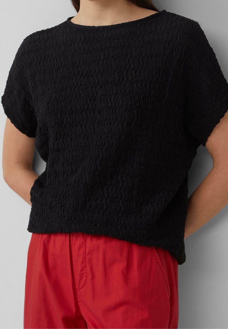 Femme portant un haut noir texturé à manches courtes et un pantalon rouge ample, debout les mains derrière le dos sur un fond gris.