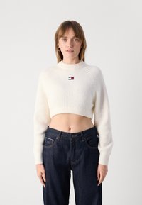 White cropped sweater with ribbed texture and high neckline, featuring a small red, white, and blue logo patch. Paired with dark denim jeans.