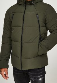 Mand i olivengrøn quilted jakke med sorte lynlåse, den ene hånd i lommen, iført sorte slidte jeans.