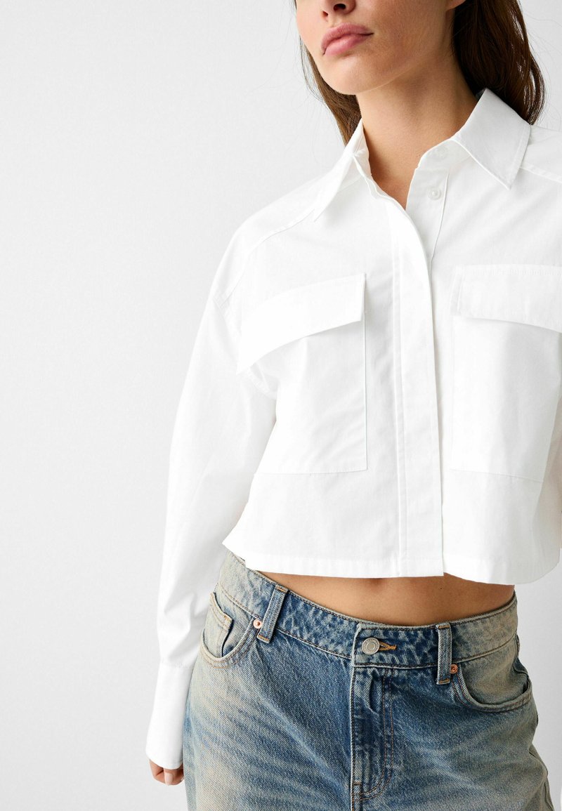 Camicia bianca cropped con collo a camicia, chiusura a bottoni e due tasche sul petto, abbinata a jeans azzurri sfilacciati.