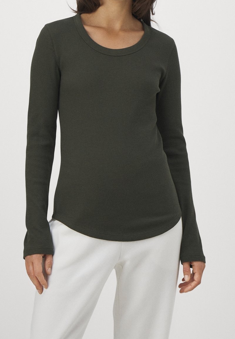 Mörkgrön, långärmad, ribbad topp med rundad halsringning och böjd nederkant. Tillverkad av ett texturerat tyg, kombinerad med vita byxor.