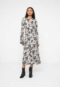 Robe midi florale en beige clair avec des motifs de fleurs noires, présentant un col en forme de trou de serrure et des manches longues, associée à des bottines noires. 