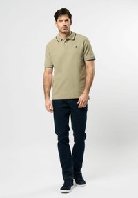 Beżowa koszulka polo z kołnierzykiem i granatowymi akcentami, zestawiona z ciemnoniebieskimi dżinsami i granatowymi sneakersami. Materiał wydaje się gładki, casualowy krój.