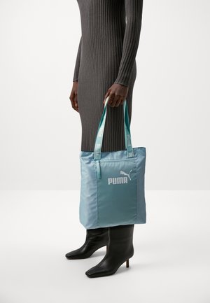 Personne en robe gris côtelée tenant un sac cabas bleu Puma avec logo, portant des bottines noires à talons sur un fond blanc.