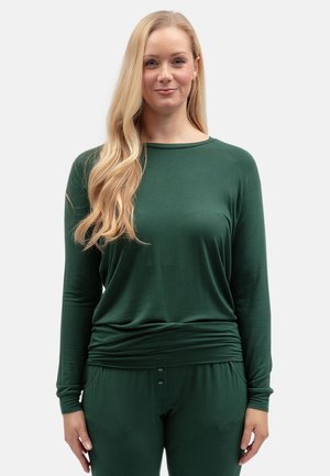 Blonde vrouw met lang golvend haar draagt een donkergroene top met lange mouwen en bijpassende broek, staand tegen een effen lichte achtergrond.