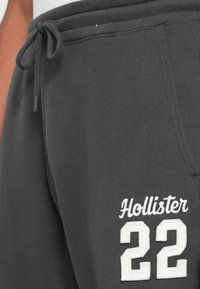 Uvećani prikaz tamnosivih trenirki s vezicom u struku, bočnom džepu i bijelom izvezenom oznakom "Hollister 22" na butini.