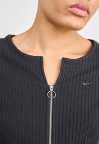 Svart ribbad zip-up topp med rund halsringning, har en silverfärgad dragkedja och en liten svart Nike-logotyp på bröstet. Strukturerad tygdetalj.