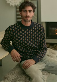 Marineblauer Pullover mit sich wiederholenden orangen Baummustern, Rundhalsausschnitt und gerippten Bündchen, getragen von einer Person, die an einem Tisch sitzt.