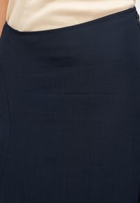 Κοντινό πλάνο ενός ατόμου που φοράει σκουρομπλε φούστα με πλαϊνό φερμουάρ και μπεζ ριμπ τοπ που είναι μαζεμένο μέσα στη φούστα.